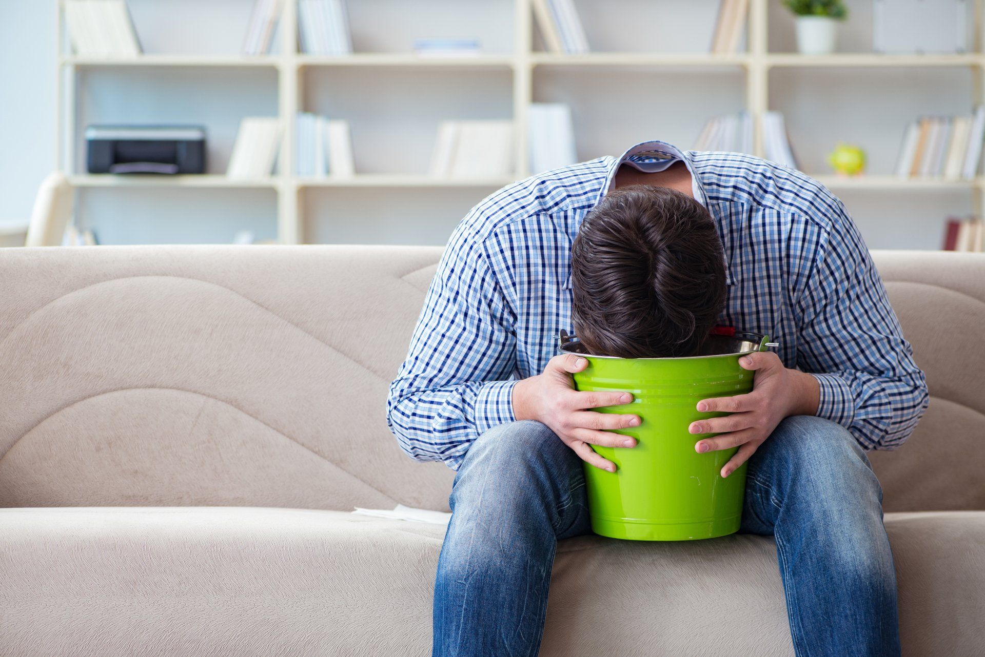 Een man zit misselijk op de bank met een emmer op schoot