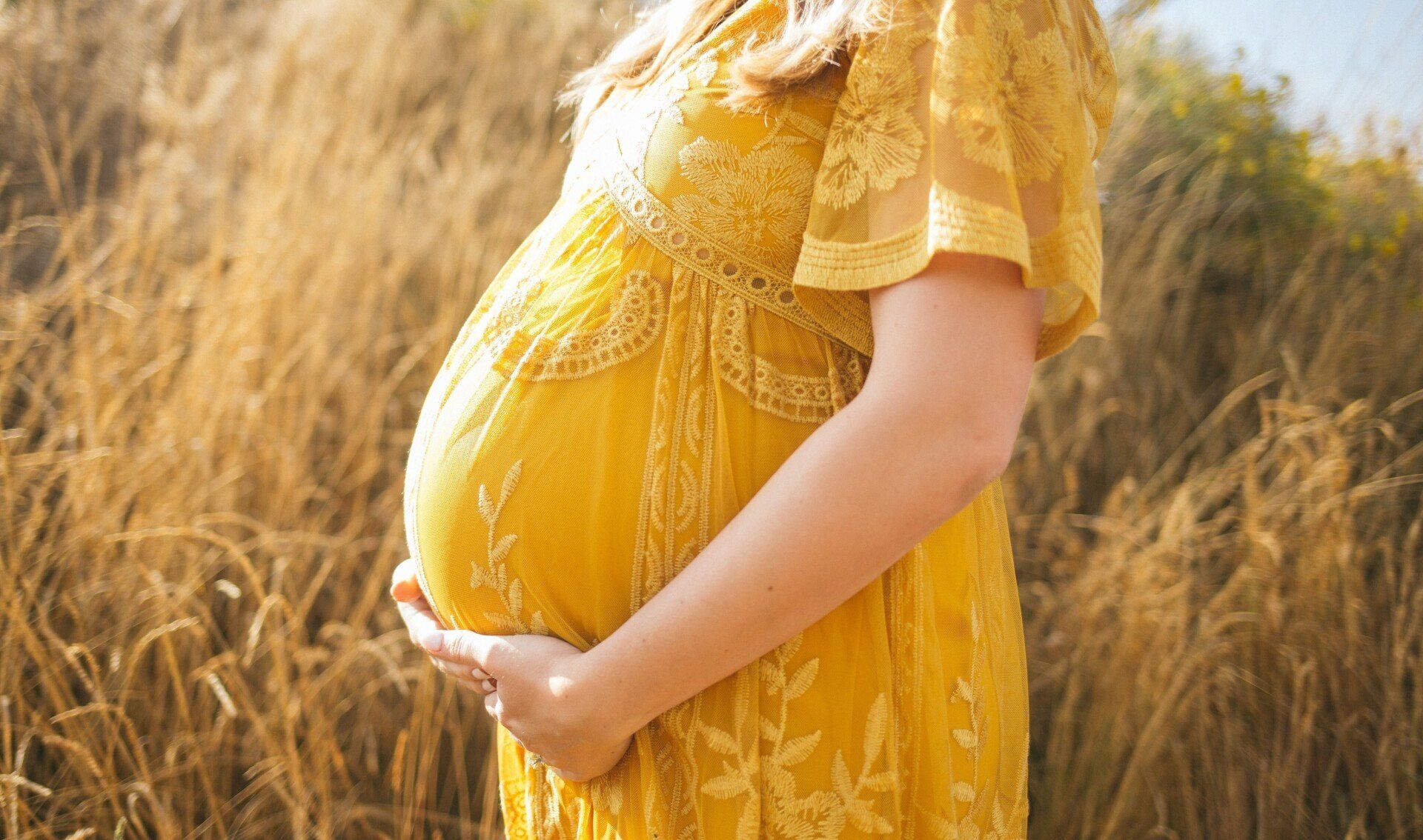 Een zwangere vrouw in een gele jurk houdt haar buik vast, in een veld